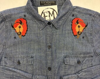 May include: Blue denim button-down shirt with two chest pockets and long sleeves. The shirt features embroidered bison heads with red and orange details on the chest. The label reads "ELLIE MONSTER NASHVILLE".