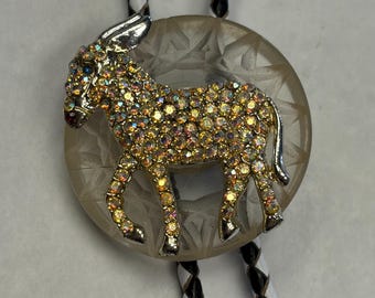 May include: A bolo tie with a gold-toned donkey pendant encrusted with rhinestones. The pendant sits on a clear, etched circular base. The tie has black and white braided cords.
