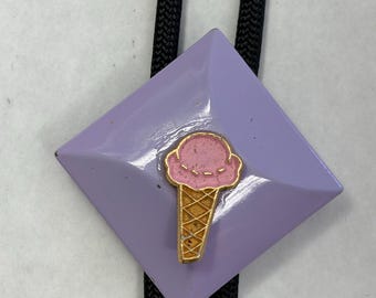 May include: A lavender bolo tie with a diamond-shaped pendant. The pendant features a gold ice cream cone with a pink scoop. The tie has a black braided cord.