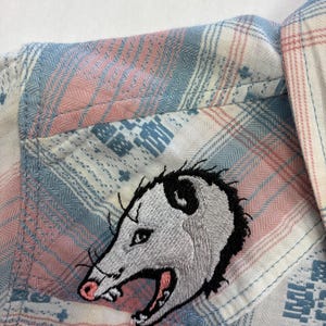 May include: A button-down shirt with a plaid pattern in shades of pink, blue, and white. An embroidered opossum patch is sewn onto the shirt pocket. The opossum has gray fur, black accents, and a pink nose and mouth.