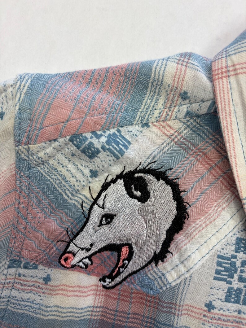 May include: A button-down shirt with a plaid pattern in shades of pink, blue, and white. An embroidered opossum patch is sewn onto the shirt pocket. The opossum has gray fur, black accents, and a pink nose and mouth.
