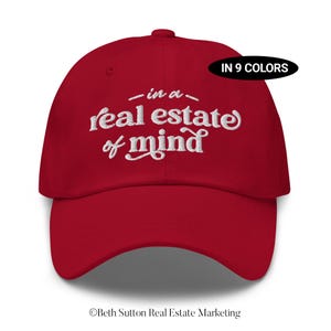 May include: A red baseball cap featuring the embroidered text "-in a- real estate of mind" in white script. The cap has a curved peak and a button on top. A black oval label reads "IN 9 COLORS".