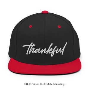 May include: Black baseball cap with a red peak and top button. The word "thankful" is embroidered in white script across the front. A stylish and comfortable accessory for everyday wear.