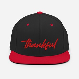 May include: A black and red baseball cap featuring the word "thankful" embroidered in red script. The cap has a flat peak and a red button on the crown.