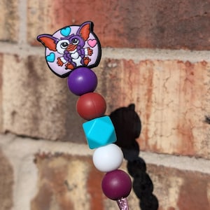 Puede incluir: Un collar de cuentas de color morado, rojo, turquesa, blanco y marrón con un dije de personaje de dibujos animados morado y rosa en la parte superior.