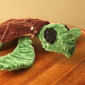 May include: A green and brown pipe cleaner turtle with black eyes. The turtle is made of pipe cleaners and has a textured surface.