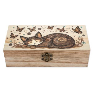 May include: Wooden box with a decorative cat and butterfly design. The cat is brown and cream-colored, with intricate patterns. Butterflies and floral accents surround the cat on the lid. The box has a metal clasp.