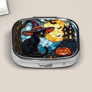 May include: A silver pill box with a Halloween-themed design. The artwork features a black cat wearing a witch's hat, a haunted house, a jack-o'-lantern, and a full moon. The background is a stained-glass style with blue and yellow hues.