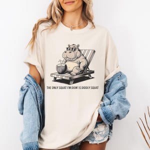 Op de afbeelding: Een wit T-shirt met een cartoonillustratie van een nijlpaard dat een zonnebril draagt en een kokosnoot vasthoudt. Het nijlpaard zit op een strandstoel. De tekst "The only squat I'm doin' is diddly squat" staat onder de afbeelding.