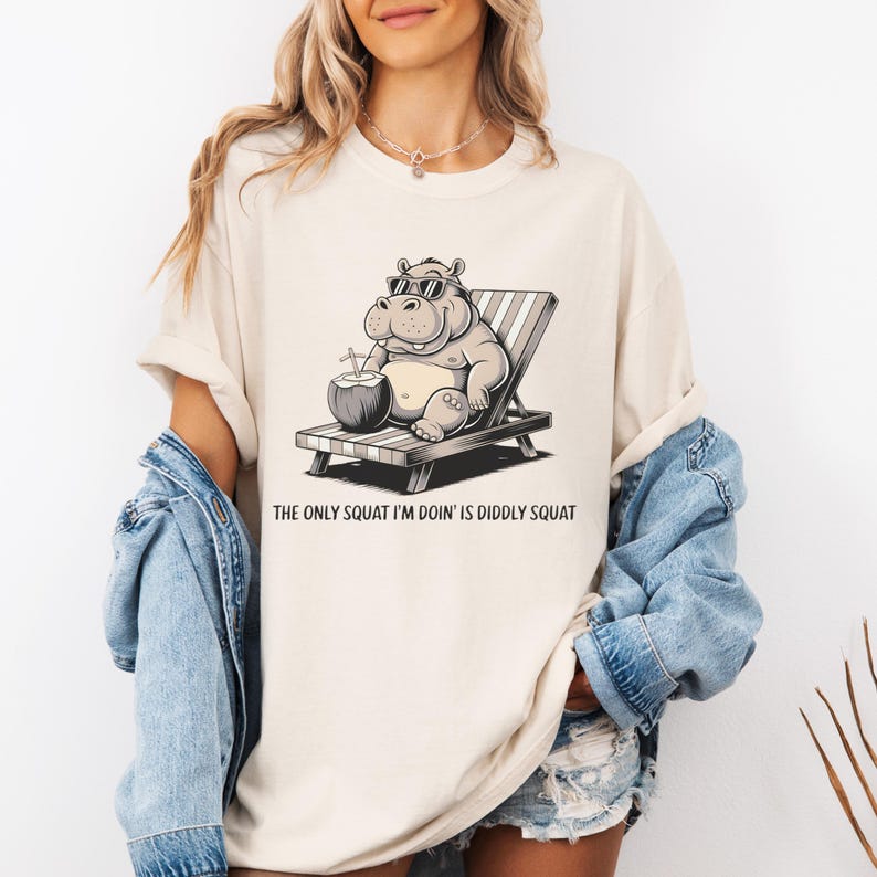 Op de afbeelding: Een wit T-shirt met een cartoonillustratie van een nijlpaard dat een zonnebril draagt en een kokosnoot vasthoudt. Het nijlpaard zit op een strandstoel. De tekst "The only squat I'm doin' is diddly squat" staat onder de afbeelding.
