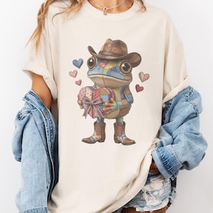 Può includere: T-shirt color crema con un personaggio di rana stravagante con cappello e stivali da cowboy, che tiene in mano una scatola regalo a forma di cuore. La rana è circondata da cuori fluttuanti nei toni del blu e del rosso. Il design ha un'estetica patchwork.