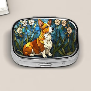 May include: A silver pill box with a stained-glass design featuring a corgi dog. The dog is brown, orange, and white, set against a blue background with white flowers and green foliage. The box has a rectangular shape.