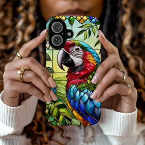 May include: A colorful phone case featuring a stained glass design of a parrot with blue, red, green, and yellow feathers. The parrot is perched on a branch with green leaves. The case has a black border and a clear camera lens.