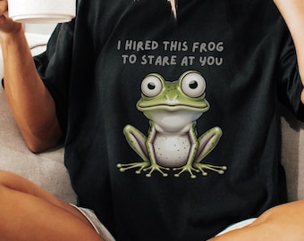 Puede incluir: Camiseta negra con una ilustración de rana de dibujos animados y el texto "I Hired This Frog To Stare At You". La rana es verde y blanca, con ojos grandes. La camiseta es de estilo casual.