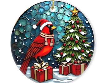 May include: A round Christmas ornament featuring a red cardinal wearing a Santa hat and scarf, standing near a decorated Christmas tree and wrapped gifts. The background is a blue and white winter scene with falling snow. A gold string is attached for hanging.