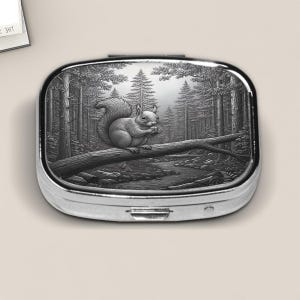 Elegant silver pill box featuring a detailed faux embossed metal illustration of a squirrel perched on a fallen log in a serene forest scene. The finely etched woodland design captures tall pine trees, soft light, and a flowing stream, giving the box a classic, storybook charm. Perfect for holding small pills, mints, or tiny keepsakes, this nature-inspired accessory makes a thoughtful gift for animal lovers and forest enthusiasts alike.
