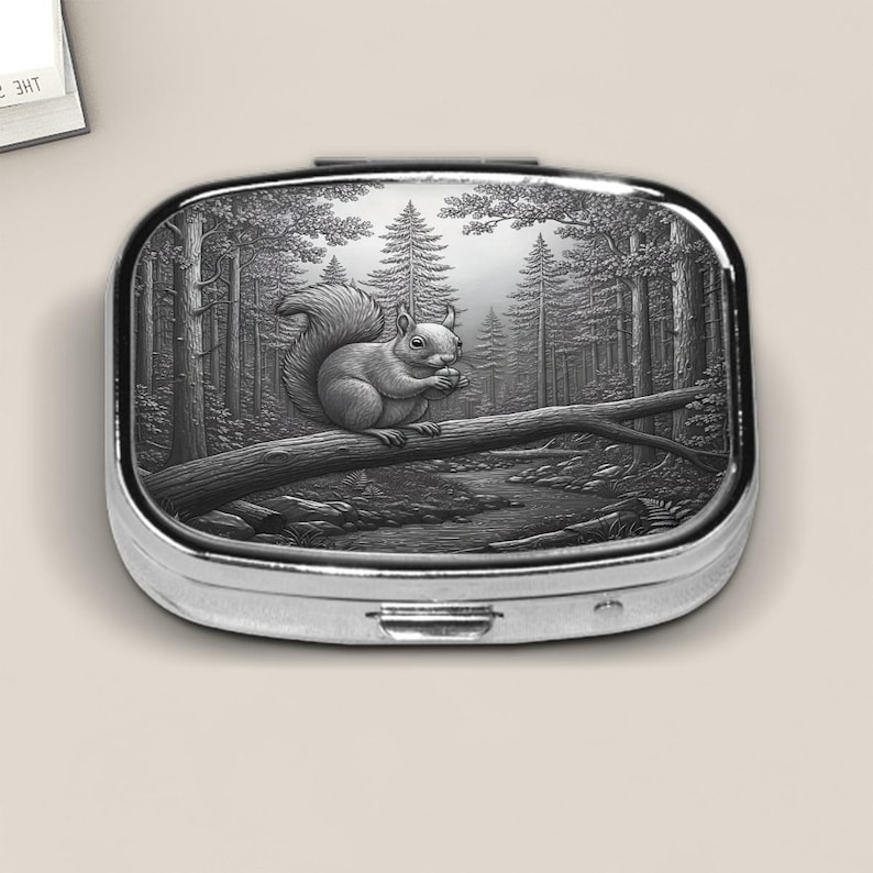 Elegant silver pill box featuring a detailed faux embossed metal illustration of a squirrel perched on a fallen log in a serene forest scene. The finely etched woodland design captures tall pine trees, soft light, and a flowing stream, giving the box a classic, storybook charm. Perfect for holding small pills, mints, or tiny keepsakes, this nature-inspired accessory makes a thoughtful gift for animal lovers and forest enthusiasts alike.