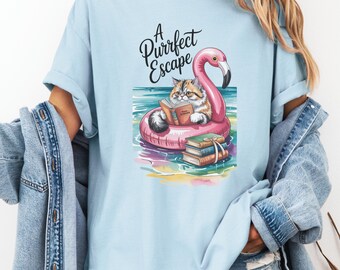 Könnte beinhalten: Hellblaues T-Shirt mit einem Aufdruck einer Katze, die ein Buch liest, während sie auf einem rosa Flamingo-Schwimmring schwimmt. Der Text "A Purrfect Escape" steht über der Katze. Mehrere Bücher sind darunter gestapelt.