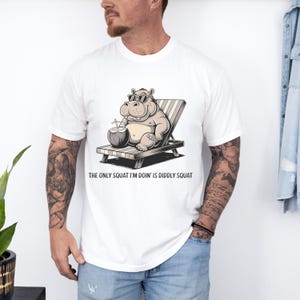 Op de afbeelding: Wit T-shirt met een cartoonillustratie van een nijlpaard met een zonnebril en een kokosnoot vasthoudend. De tekst "The only squat I'm doin' is diddly squat" staat onder de afbeelding.