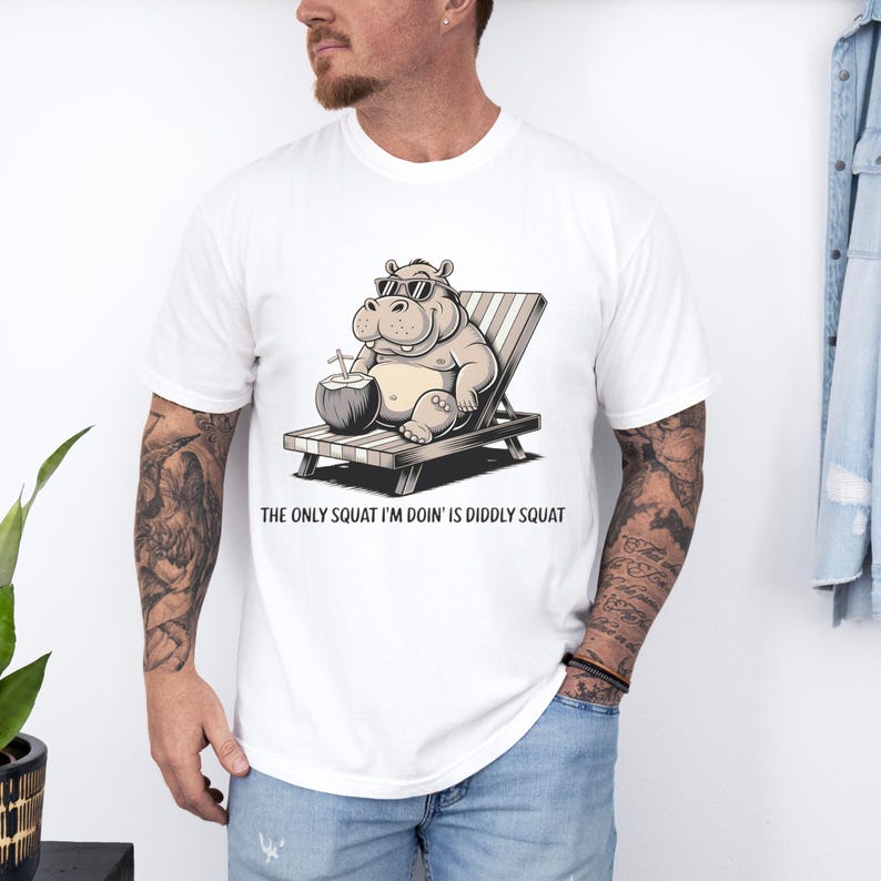Op de afbeelding: Wit T-shirt met een cartoonillustratie van een nijlpaard met een zonnebril en een kokosnoot vasthoudend. De tekst "The only squat I'm doin' is diddly squat" staat onder de afbeelding.