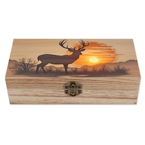 May include: Wooden box with a deer silhouette against a sunset backdrop. The box features a hinged lid and a metal clasp. The artwork depicts a deer standing in front of a sunset with mountains and trees. The box has a natural wood grain finish.