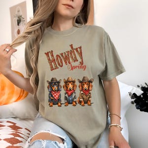 May include: A light green t-shirt with the text "Howdy Spring" and a graphic of three dachshund dogs wearing cowboy hats and bandanas.