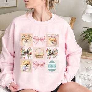 Op de afbeelding: Een lichtroze sweatshirt met een raster van Pasen-thema illustraties. Het ontwerp bevat dwergkeeshonden met bloemenkransen en konijnenoren, strikken, een mand met eieren en een versierd ei. De afbeeldingen zijn in pastelkleuren.