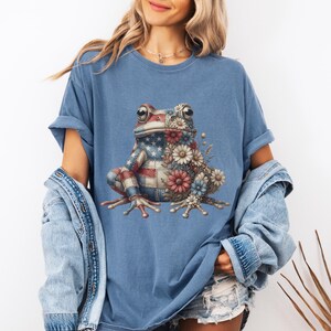 Könnte beinhalten: Blaues T-Shirt mit einem Frosch-Design. Der Frosch ist mit einem amerikanischen Flaggenmuster und Blumen in Rot, Weiß und Blau verziert. Das T-Shirt hat kurze Ärmel und eine lockere Passform.