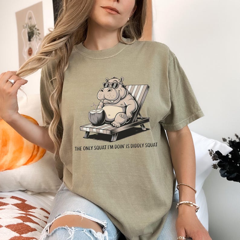 Op de afbeelding: Een lichtgroen T-shirt met een cartoonillustratie van een nijlpaard met een zonnebril en een kokosnoot in zijn hand. De tekst "The only squat I'm doin' is diddly squat" staat op het T-shirt gedrukt.
