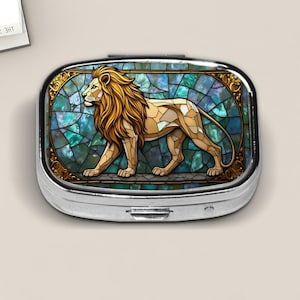May include: A silver pill box with a stained-glass design featuring a golden lion. The lion is depicted in a walking pose against a backdrop of teal and blue mosaic tiles, framed by a gold border.