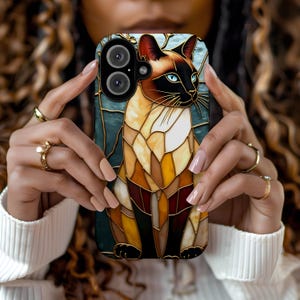 Puede incluir: Una funda de teléfono de vidrio coloreado con un gato siamés de ojos azules. El gato está representado de pie con un diseño detallado y colorido.
