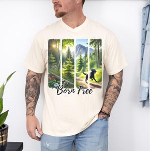Puede incluir: Una camiseta de color crema con cuello en V y un estampado gráfico de una escena de bosque con un perro negro corriendo por un camino. El texto "Born Free" está impreso debajo del gráfico.
