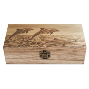May include: Wooden box with a hinged lid and a metal clasp. The box features a detailed carved relief of three dolphins leaping out of ocean waves. The wood has a natural grain and a light brown color.