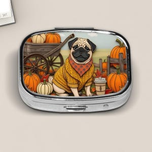 May include: A silver pill box featuring a cartoon pug wearing a yellow sweater and a plaid scarf, set in a fall scene with pumpkins, a wooden cart, and a fence. A coffee cup is also visible.