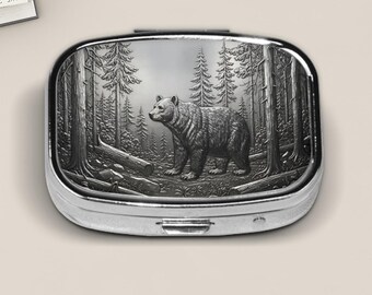 May include: Silver-colored pill box with a detailed forest scene and a bear. The bear is walking through a forest of tall trees. The pill box has a rectangular shape with rounded edges and a hinged lid.