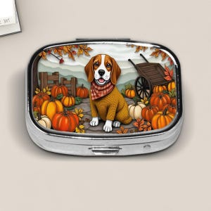 May include: A silver pill box featuring a beagle dog wearing a sweater and a plaid scarf, surrounded by pumpkins, fall leaves, and a wooden cart. The dog is sitting in a pumpkin patch. The background includes a wooden fence and a mountain range.