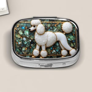 May include: A silver pill box with a white poodle design. The poodle is set against a background of green and gold mosaic. The dog has fluffy white fur and is in a standing pose. The box is rectangular with rounded edges.