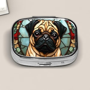 May include: A silver pill box with a stained-glass design featuring a pug's face. The pug has a tan coat, black ears and muzzle, and large brown eyes. The background is light blue with red and green accents.