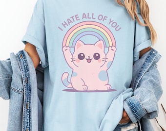 Può includere: T-shirt azzurra con un gatto rosa con macchie blu che tiene un arcobaleno. Il testo "I HATE ALL OF YOU" è sopra. La maglietta ha maniche corte e una vestibilità ampia.