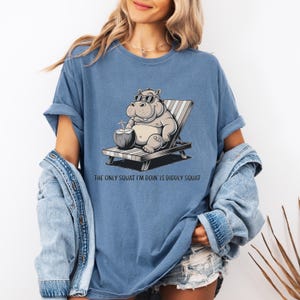 Op de afbeelding: Een blauw T-shirt met een cartoonillustratie van een nijlpaard dat een zonnebril draagt en ontspant op een strandstoel met een kokosnootdrankje. De tekst "The only squat I'm doin' is diddly squat" staat onder de afbeelding.
