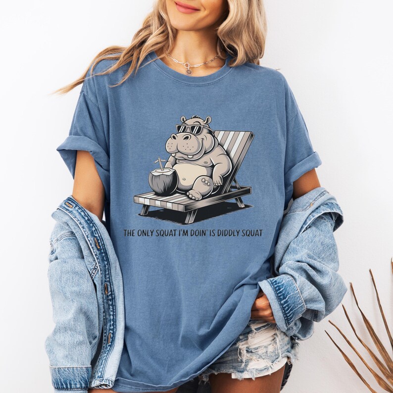 Op de afbeelding: Een blauw T-shirt met een cartoonillustratie van een nijlpaard dat een zonnebril draagt en ontspant op een strandstoel met een kokosnootdrankje. De tekst "The only squat I'm doin' is diddly squat" staat onder de afbeelding.