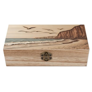 May include: A wooden box with a carved image of a beach scene with seagulls flying over the ocean and a cliff in the background. The box has a brass latch.