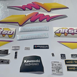 May include: A collection of adhesive decals for a Kawasaki AR50 motorcycle, featuring large yellow and pink graphic accents, AR50 logos, Kawasaki text, uni-trak labels, and various small safety and technical warning stickers with black and white text.