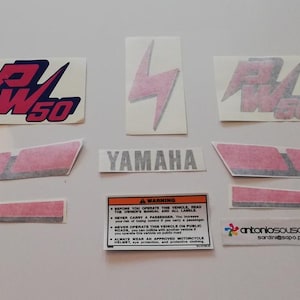 Puede incluir: Un conjunto de pegatinas para motocicleta PW50 que incluye dos logotipos PW50, un rayo, la marca Yamaha, dos franjas laterales rosas largas, dos franjas rosas más cortas, una etiqueta de advertencia de seguridad y una pequeña pegatina rectangular.