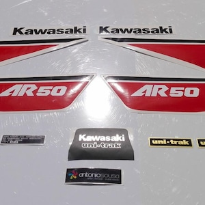 Può includere: Un set di adesivi per moto Kawasaki AR50 con grafiche rosse, nere e bianche. La collezione include due grandi adesivi per pannelli laterali con testo AR50, due loghi Kawasaki, due etichette Uni-Trak e ulteriori piccoli adesivi identificativi.