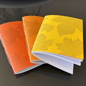 Könnte beinhalten: Drei kleine Notizhefte mit Papierumschlägen in Orange-, Bernstein- und Gelbtönen. Jeder Umschlag zeigt ein wiederkehrendes Muster aus Ahornblattsilhouetten. Die Hefte sind mit einer sichtbaren Naht am Rücken gebunden und enthalten weiße linierte Seiten.