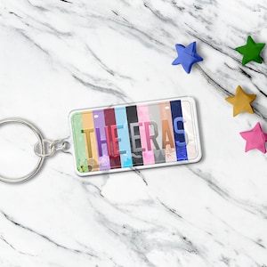 May include: A rectangular keychain with the text "THE ERAS" in a colorful, vertical stripe design. The keychain has a silver key ring and is surrounded by colorful star-shaped decorations.