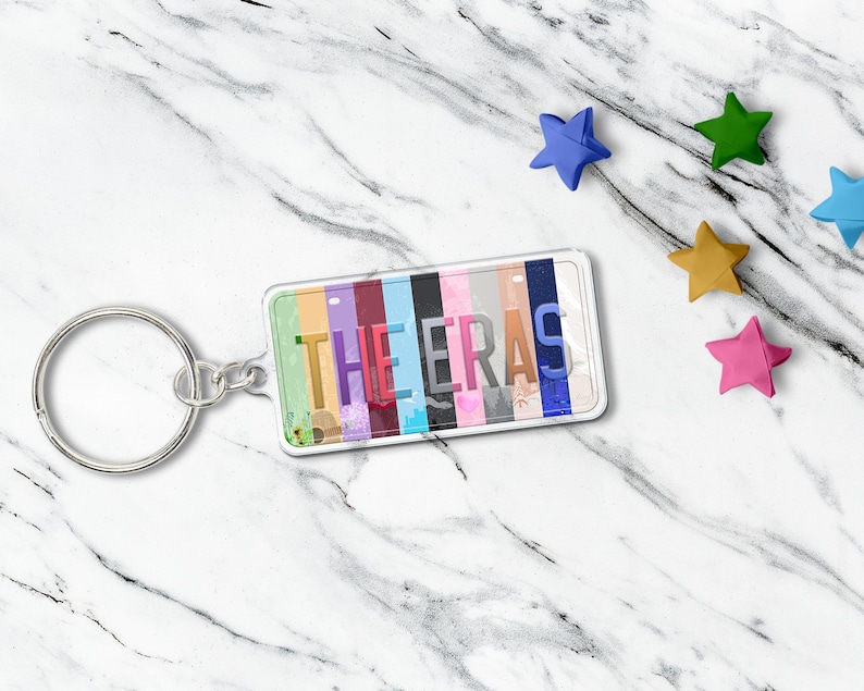 May include: A rectangular keychain with the text "THE ERAS" in a colorful, vertical stripe design. The keychain has a silver key ring and is surrounded by colorful star-shaped decorations.