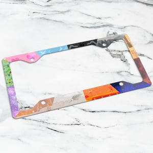 May include: A colorful license plate frame with a variety of patterns and colors. The frame features sections of pink, blue, black, orange, and purple, each with unique designs including birds, flowers, and abstract shapes. The frame is designed to fit a standard license plate.