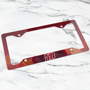 Peut inclure: Un cadre de plaque d'immatriculation rectangulaire présentant un dégradé de tons rouge foncé et orange avec un motif de feuilles d'automne. Le mot RED est imprimé en lettres capitales rose clair sur la barre horizontale inférieure du cadre.